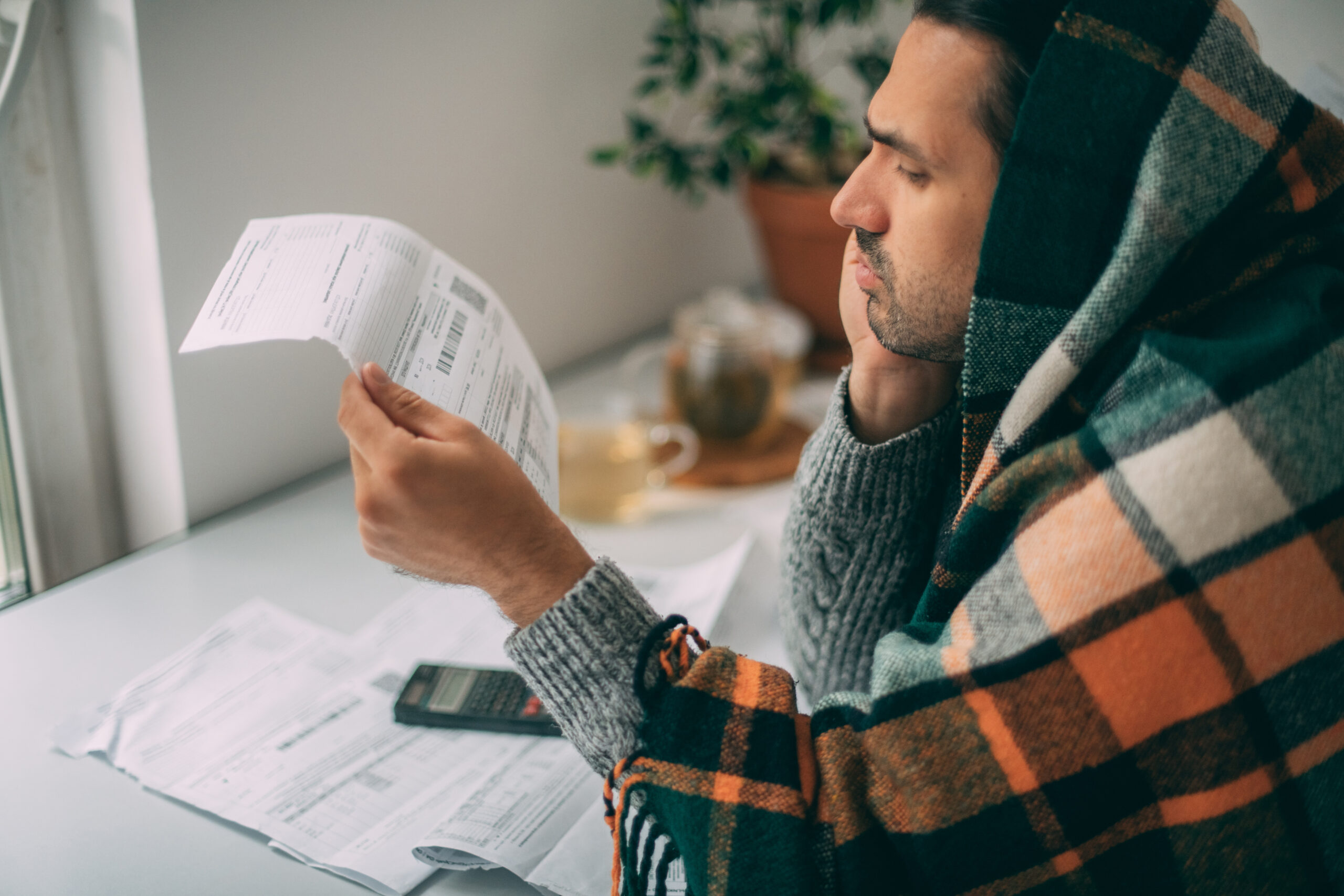 A sad man in a warm plaid is sitting at a table with utility bills in his hands.