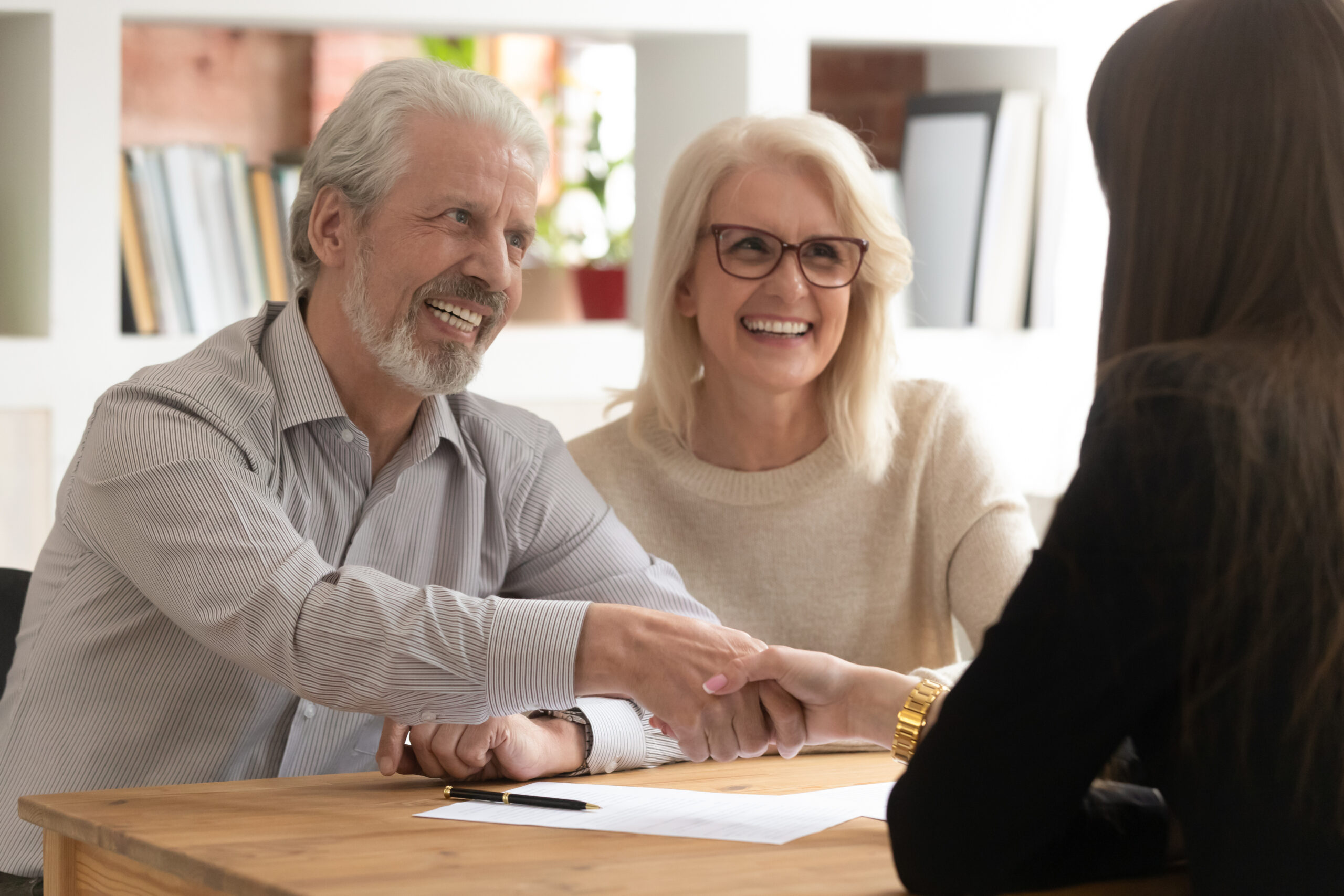 People sit at desk in office focus on aged couple handshaking with female realtor real estate agent succeed agreement, after sign contract client express gratitude shake hands, successful deal concept